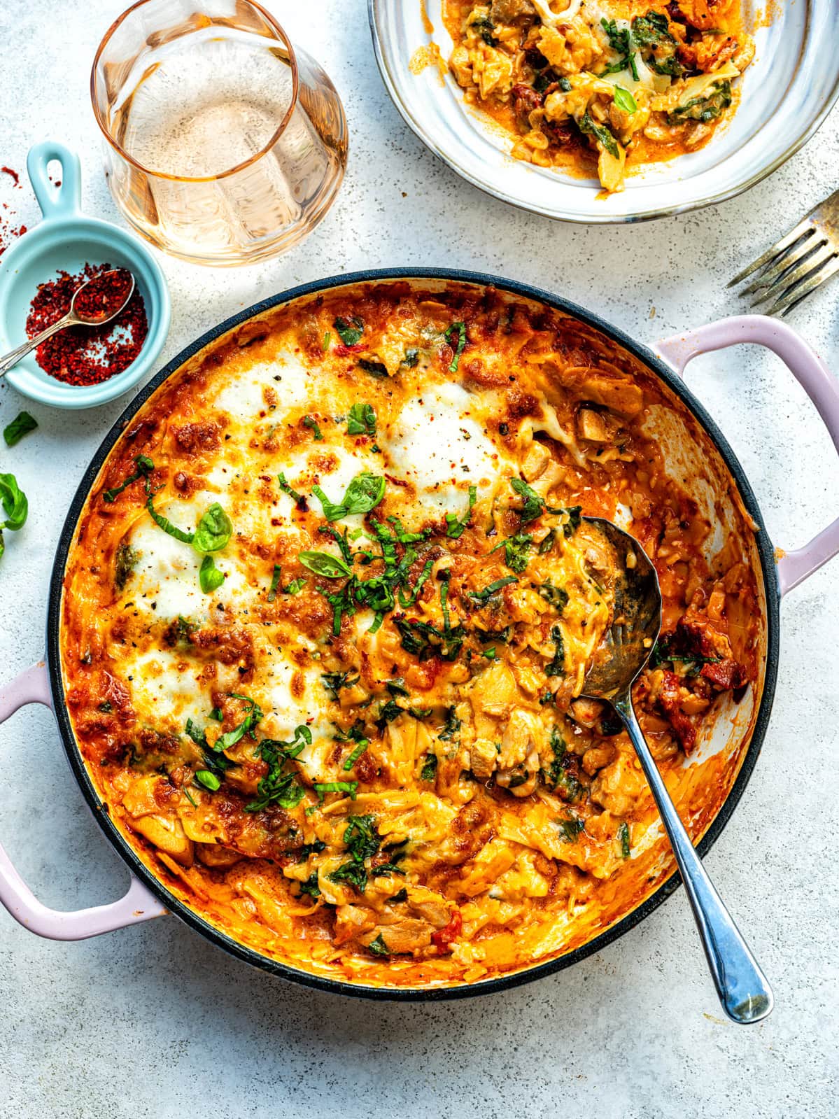 A round pan filled with cheesy Marry Me Skillet Lasagna, topped with fresh herbs, sits on a light surface. A spoon rests in the pan, a serving is plated nearby, and a glass of water and small dish of red spices are beside the pan.
