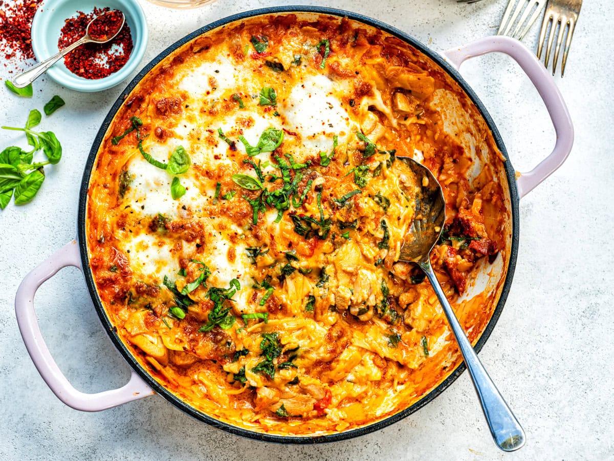 A round baking dish filled with baked pasta in a creamy tomato sauce, reminiscent of marry me chicken lasagna, topped with melted cheese and fresh basil. A serving spoon rests inside, with a small bowl of red pepper flakes and herbs beside the dish.