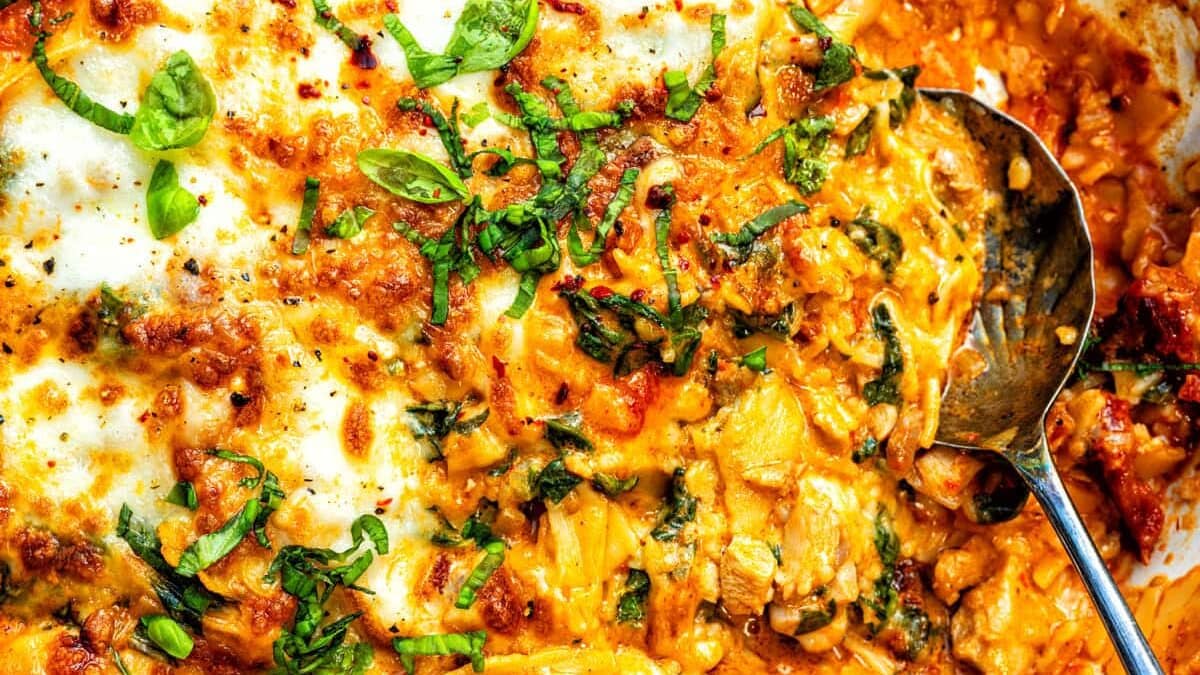 Close-up of a cheesy, baked broken marry me chicken lasagna with melted cheese, tomato sauce, ground meat, and fresh herbs like basil and parsley, all made in one pan with a metal serving spoon resting on the side.