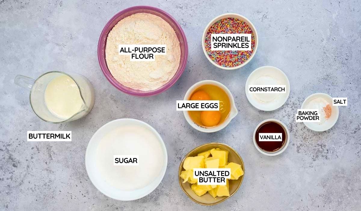 A top-down view of baking ingredients in bowls and cups, labeled for a Dot Cake Recipe: all-purpose flour, sugar, buttermilk, large eggs, unsalted butter, nonpareil sprinkles—perfect for making delightful Dot Cakes.