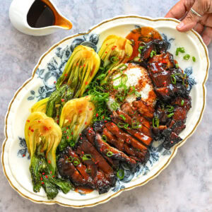 A hand holds a platter with air fryer char siu pork—glazed, sliced meat—alongside steamed bok choy, rice, and green onions. A small pitcher of sauce is beside the platter on a marble surface.