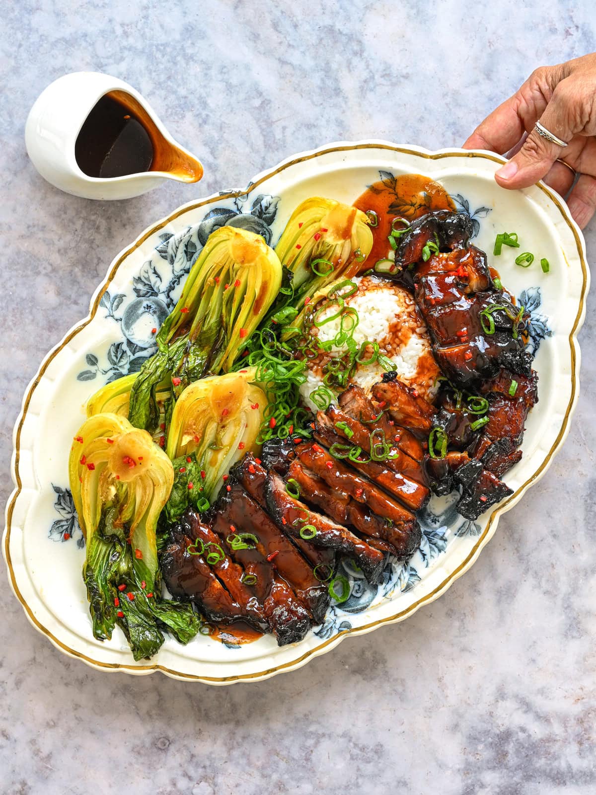 A hand holds a platter with air fryer char siu pork—glazed, sliced meat—alongside steamed bok choy, rice, and green onions. A small pitcher of sauce is beside the platter on a marble surface.