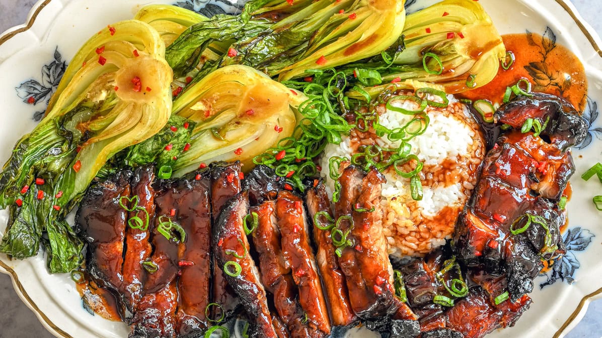 Char siu grilled glazed meat slices with steamed bok choy, white rice, and chopped green onions, all served on a decorative plate with a drizzle of sauce.