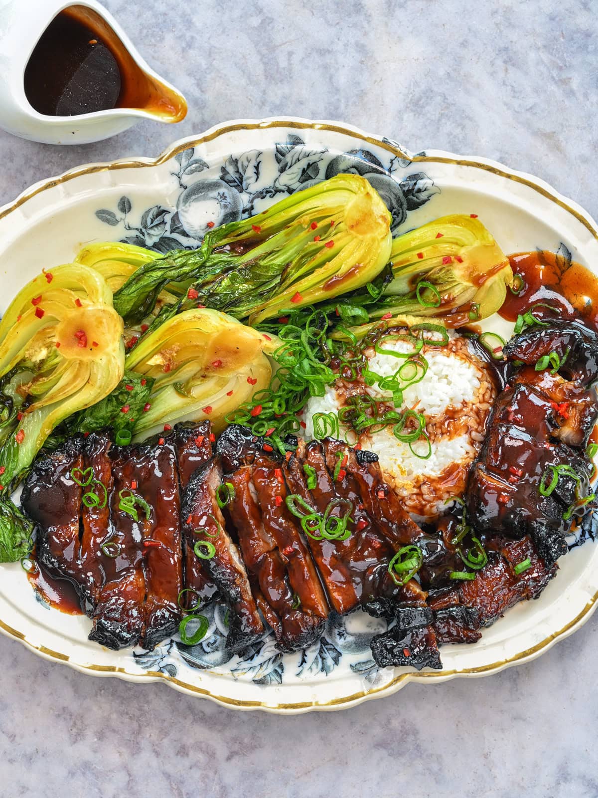 Sliced glazed meat with rice, bok choy, and chopped green onions on a floral plate. A small pitcher of sauce sits nearby.