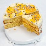 A layered whole lemon cake with white frosting, topped with yellow candied fruit slices, small white flowers, and purple sprinkles. A large slice of this delightful boiled lemon cake is cut out and placed slightly apart on a white cake stand.