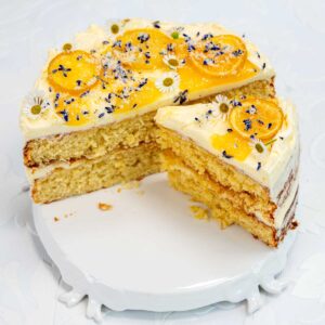 A layered whole lemon cake with white frosting, topped with yellow candied fruit slices, small white flowers, and purple sprinkles. A large slice of this delightful boiled lemon cake is cut out and placed slightly apart on a white cake stand.