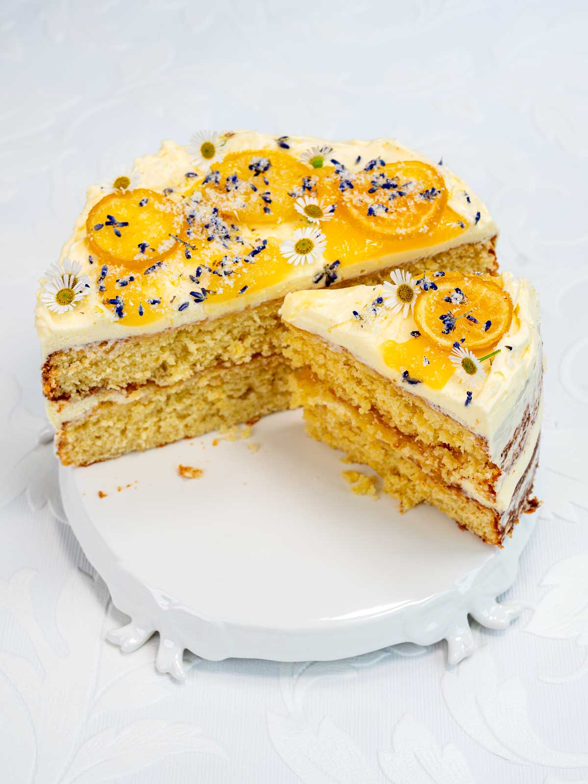A frosted layer cake decorated with yellow fruit slices, purple flower petals, and small white flowers, with one slice cut and slightly separated on a white cake stand.