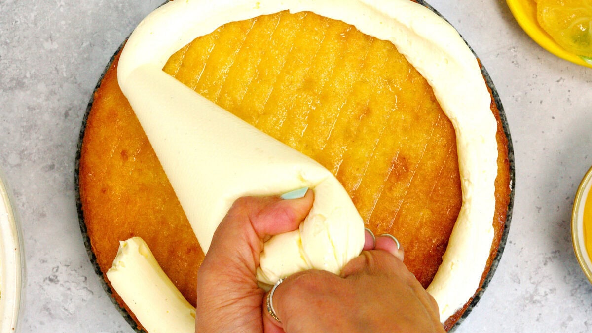 A person pipes a thick ring of white frosting around the edge of a yellow cake layer on a gray surface.