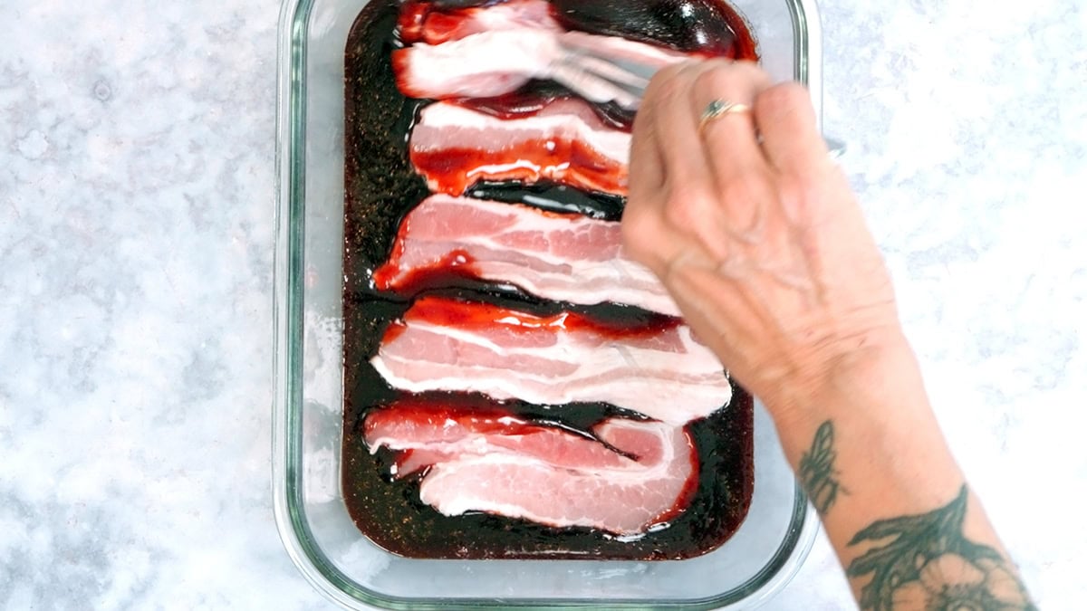 A hand with a tattoo uses a fork to coat raw bacon slices in a dark red marinade inside a glass baking dish on a light-colored countertop.