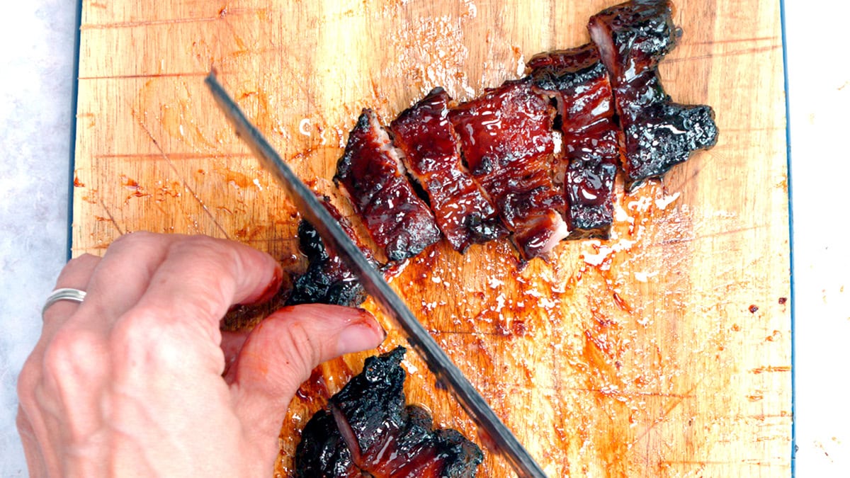 A hand slices glazed, Chinese BBQ pork belly slices on a wooden cutting board with a large knife. The meat is juicy and caramelized, and the board is stained with sauce.