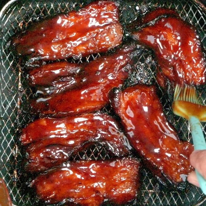 Barbecue-glazed ribs being brushed with sauce inside an air fryer basket, with a hand holding a brush applying more sauce to the ribs.