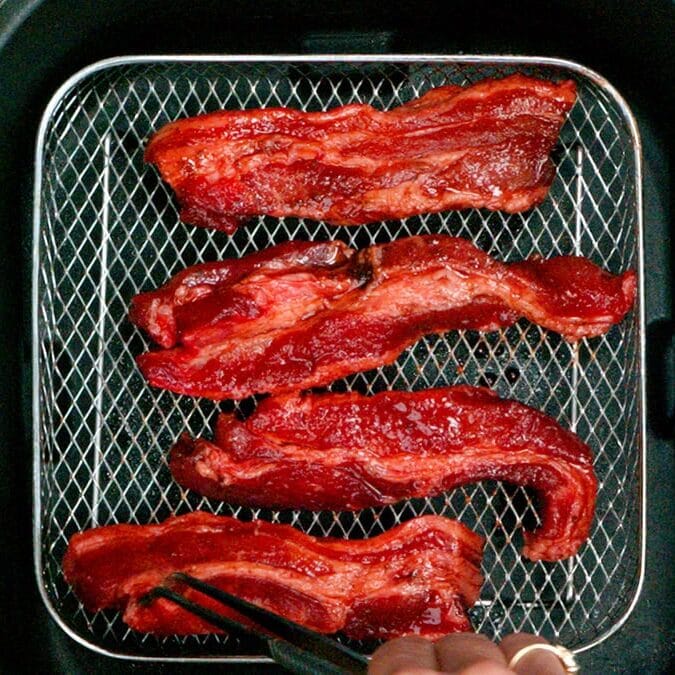 Four pieces of seasoned meat are placed inside an air fryer basket. A hand at the bottom right uses tongs to arrange one piece. The meat appears red and uncooked, resting on a wire rack.