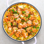 A round casserole dish with cooked salmon chunks, rice, corn, green onions, and black sesame seeds, garnished with red pepper flakes.