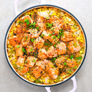A round casserole dish with cooked salmon chunks, rice, corn, green onions, and black sesame seeds, garnished with red pepper flakes.