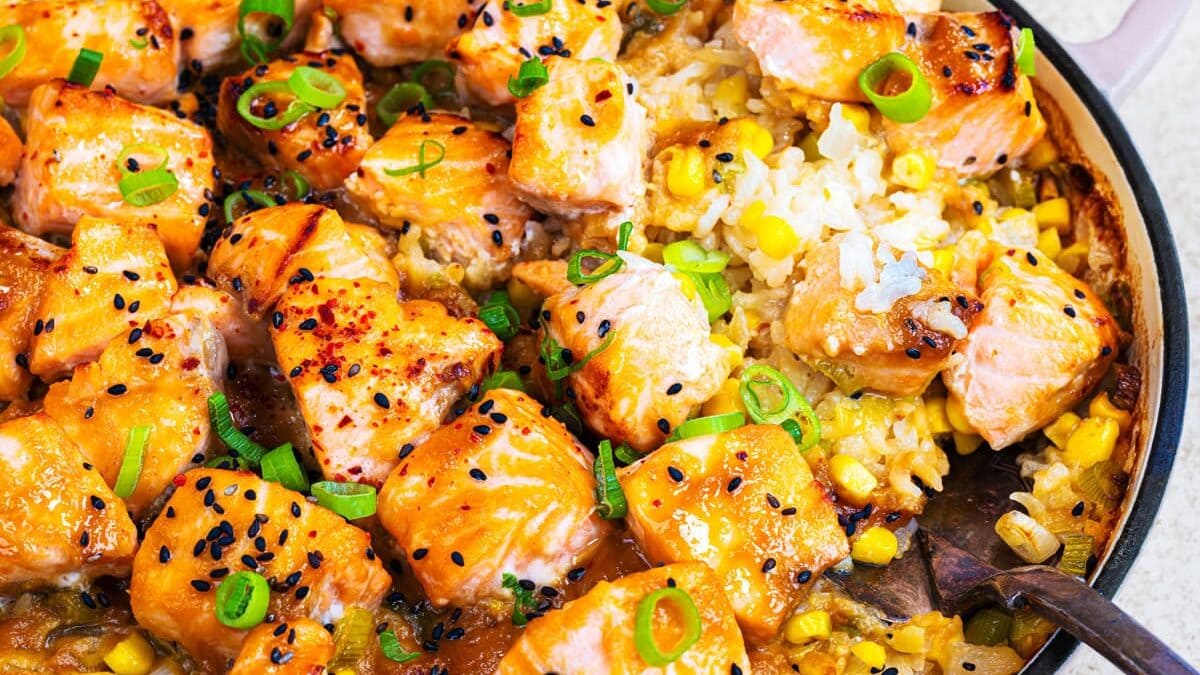 A pan filled with cooked salmon chunks on top of creamy rice with corn, garnished with sliced green onions and black sesame seeds, with a serving spoon on the side.