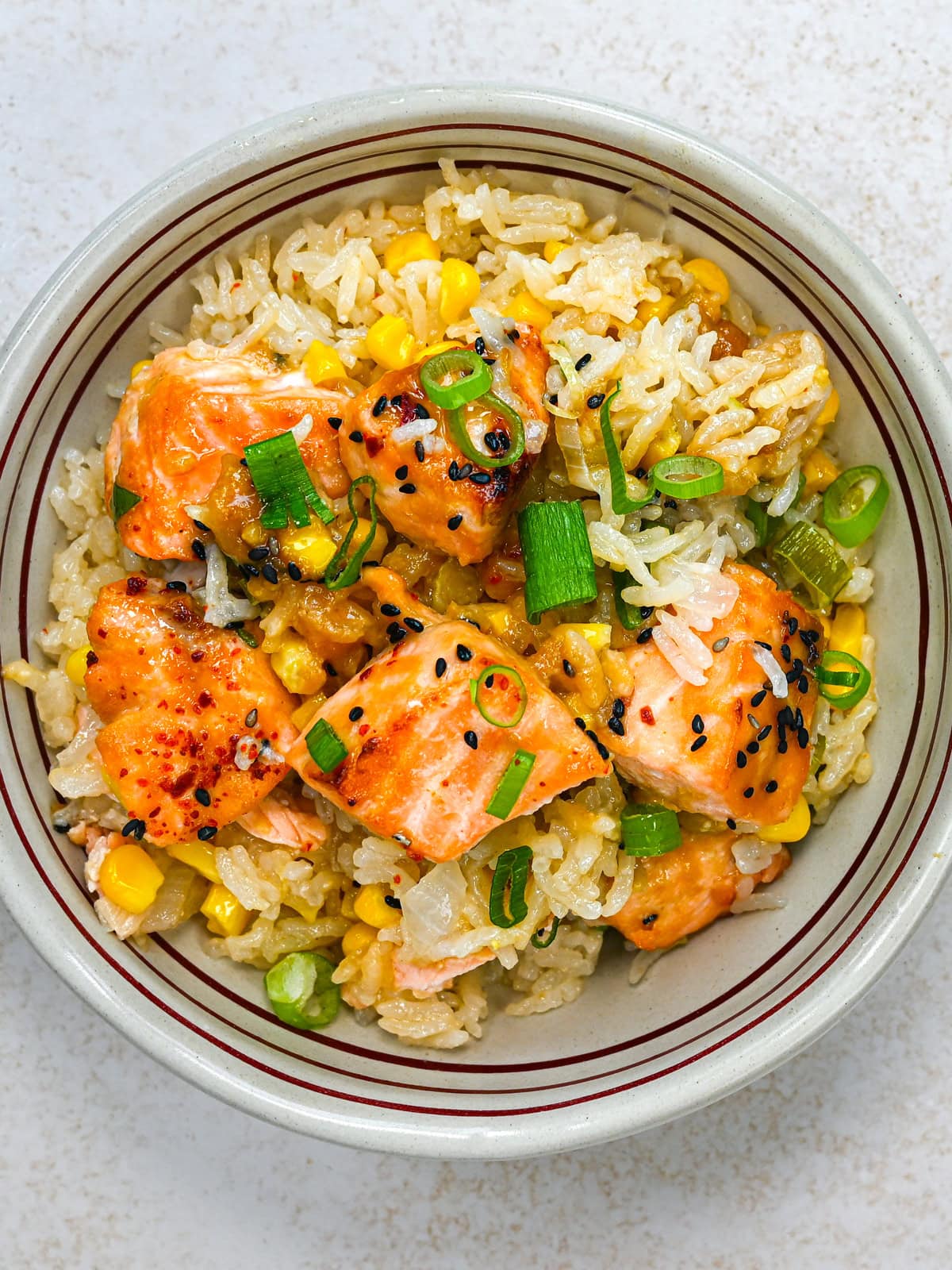 A bowl of coconut rice topped with cooked salmon, corn, chopped green onions, and black sesame seeds, served on a light background.