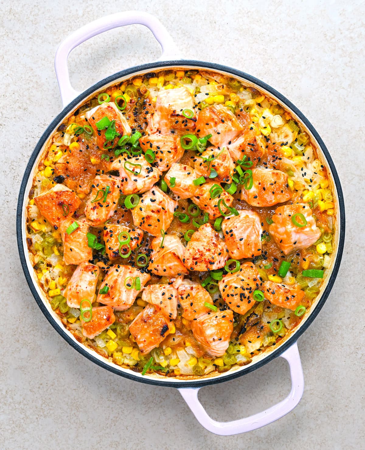 A round casserole dish with cooked salmon chunks, rice, corn, green onions, and black sesame seeds, garnished with red pepper flakes.