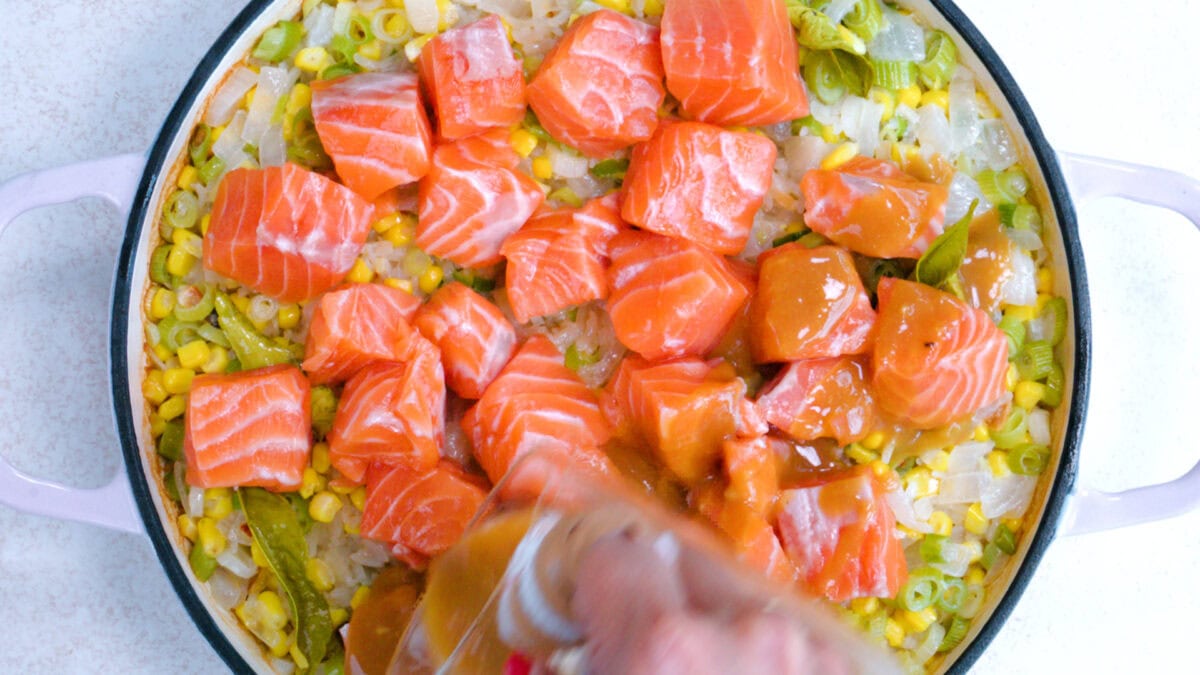Cubes of raw salmon, green onions, corn, and rice in a round pan, with a hand pouring a brown sauce over the ingredients.
