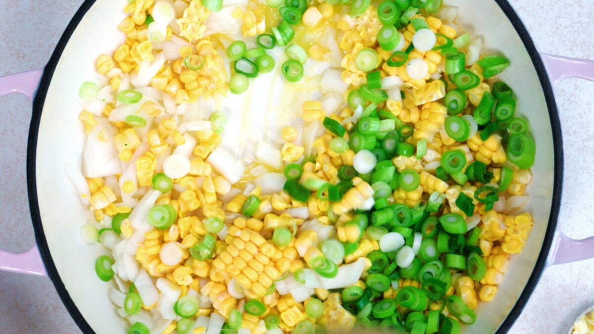 Chopped green onions, corn kernels, and sliced white onions are in a light-colored pot, ready to be cooked. The ingredients are fresh and evenly distributed in the pot.