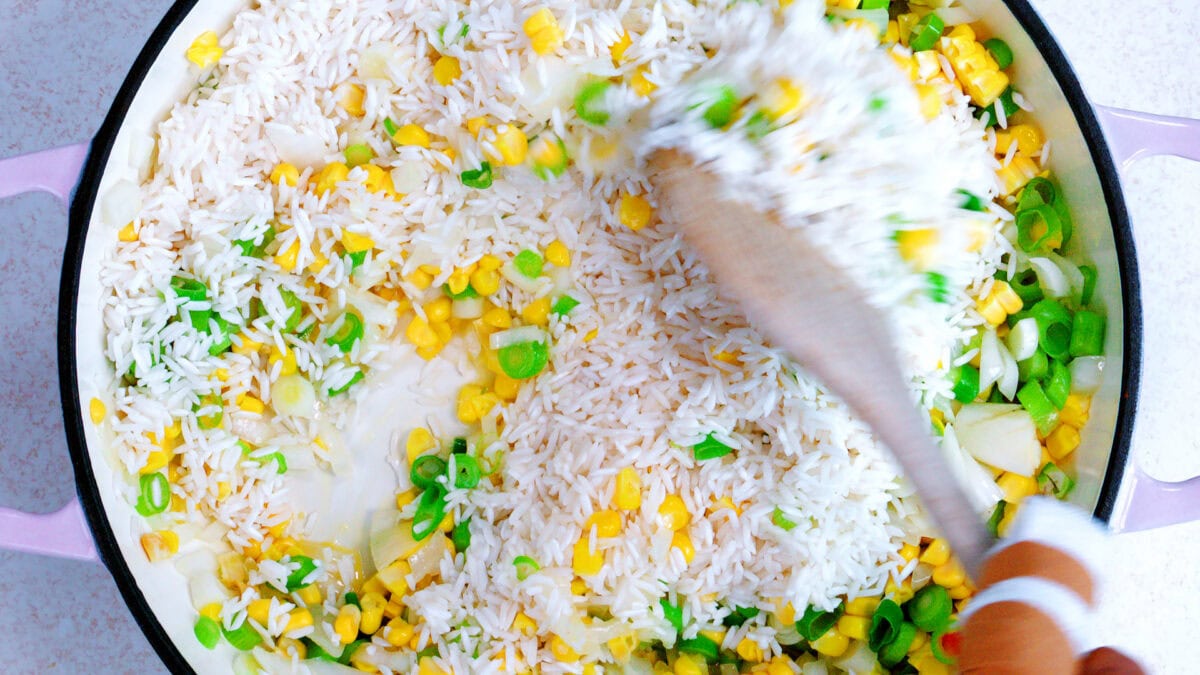 A close-up of a skillet with rice, corn, and chopped green onions being stirred with a wooden spoon.