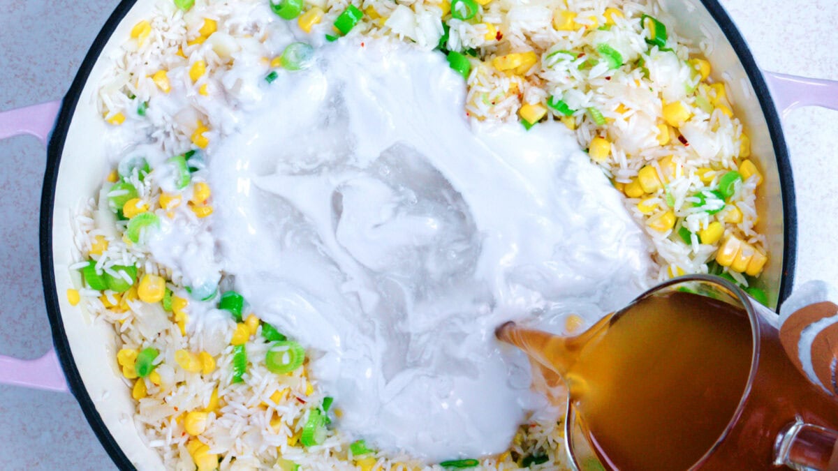 A pot of fried rice with corn, peas, and green onions is being mixed with coconut milk and broth, poured from a jug.