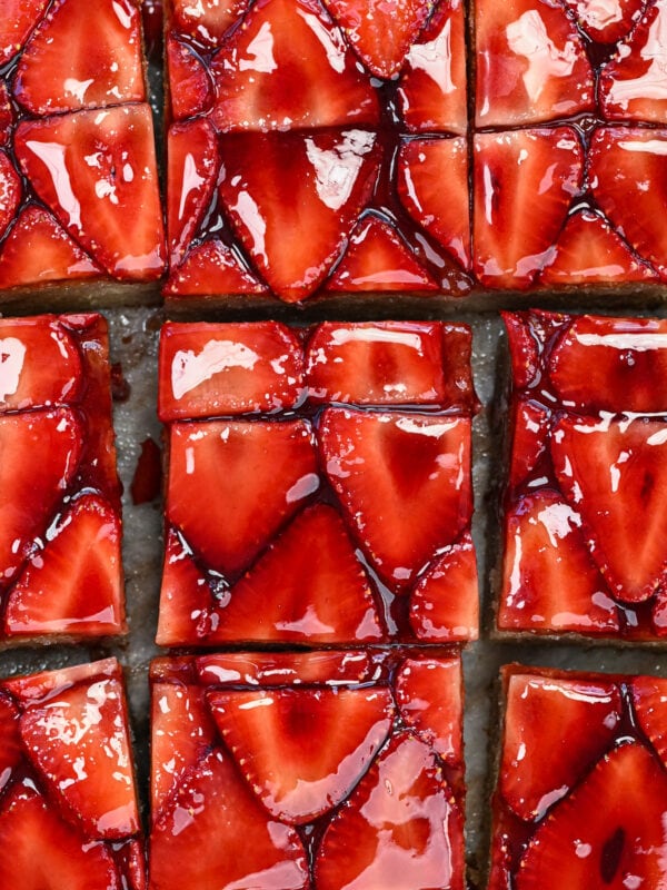 Close-up of glossy strawberry cake slices, arranged in a grid pattern. The strawberries are thinly sliced and coated with a shiny glaze, covering a pastry base.