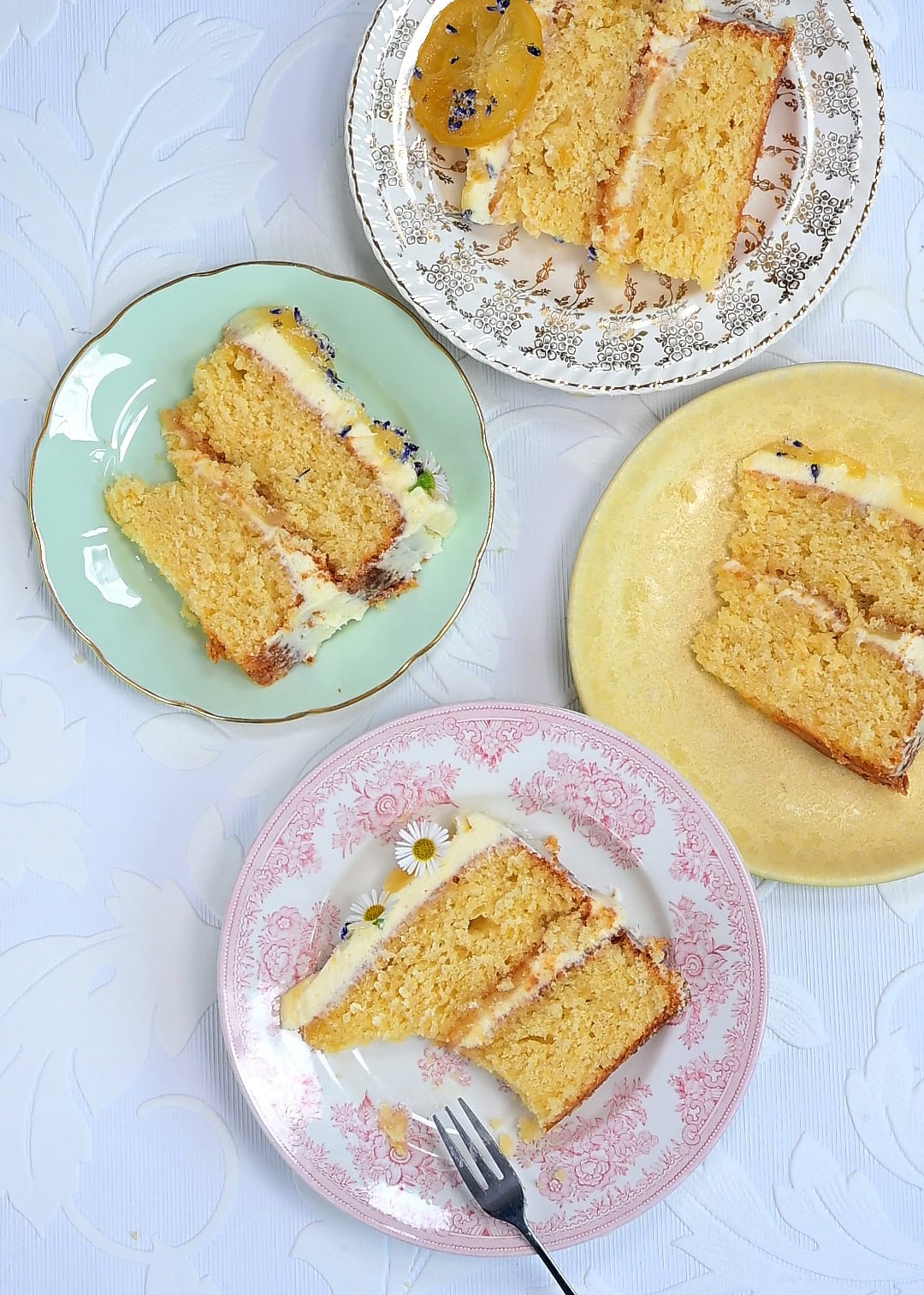 Four vintage plates with slices of yellow whole lemon layer cake and frosting are arranged on a white patterned tablecloth. One slice is garnished with edible flowers, and another has a small fork on the plate.