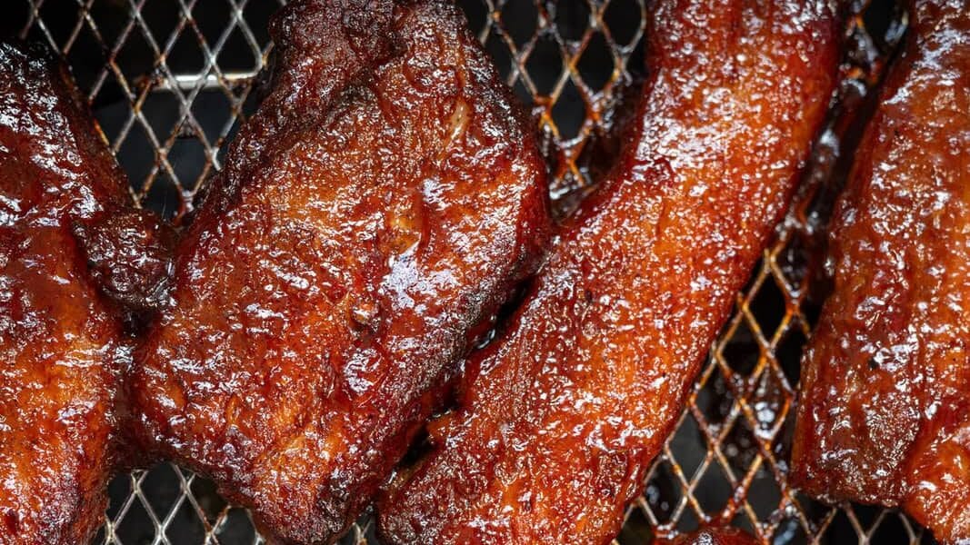 Barbecue pork strips with a glossy, caramelized glaze are arranged on a metal grill grate, their rich reddish-brown color indicating they are freshly cooked and ready to eat.