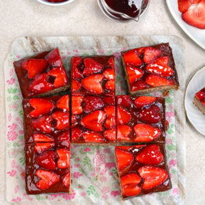 Seven square slices of glazed strawberry cake are arranged on floral parchment paper, surrounded by a bowl of fresh strawberries, a small jug of red sauce, and a plate with extra strawberries on a light surface.