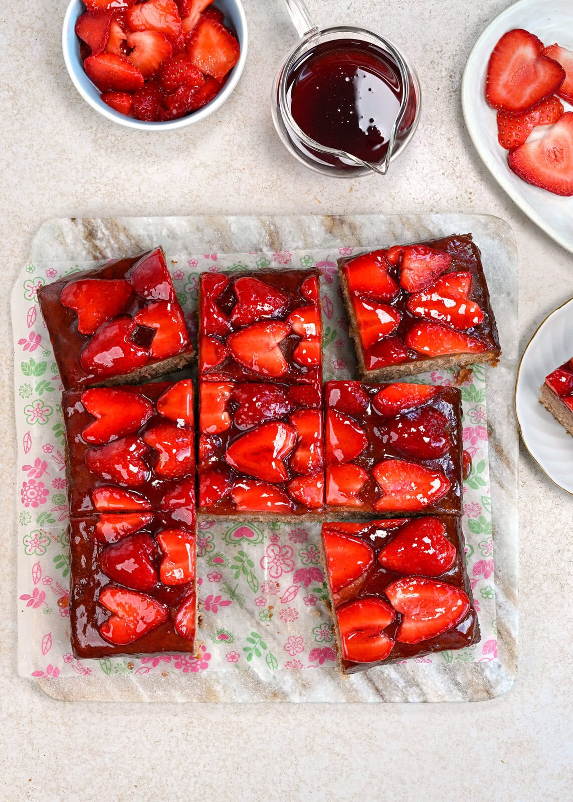 Seven square slices of glazed strawberry cake are arranged on floral parchment paper, surrounded by a bowl of fresh strawberries, a small jug of red sauce, and a plate with extra strawberries on a light surface.
