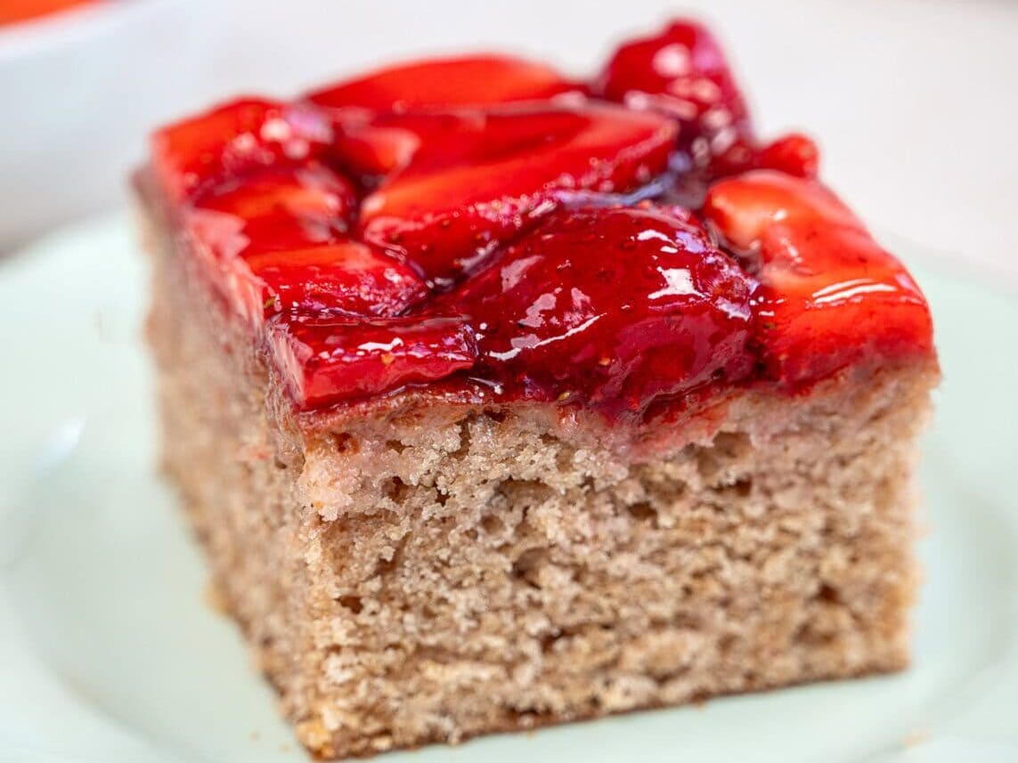 A slice of cake topped with glazed strawberries sits on a light green plate. The cake appears moist and fluffy, with a layer of shiny red fruit on top. In the background, there is a blurry bowl of sliced strawberries.