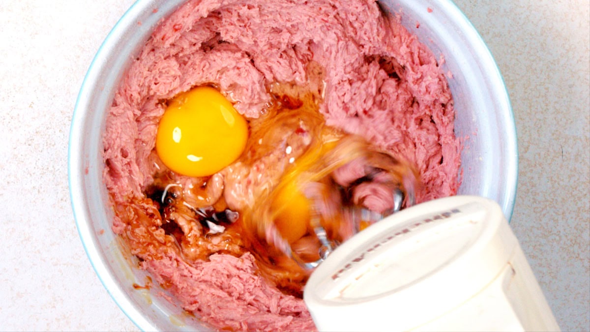 A hand mixer blends an egg and vanilla into a bowl of pink strawberry cake batter, creating a swirling motion.