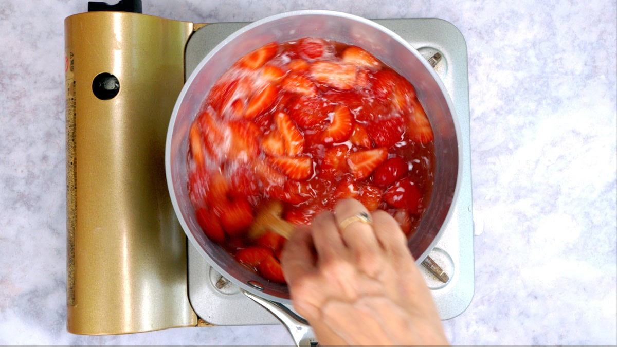 A hand stirs cut strawberries cooking in a pot on a portable stove, seen from above. The fruit mixture appears to be simmering, with some liquid bubbling around the strawberries.