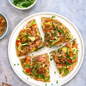 A white plate with four slices of sushi pizza, with crispy rice paper base topped with raw fish, avocado, sauce, and chopped chives, surrounded by bowls of crispy onions and sliced cucumber on a gray surface.