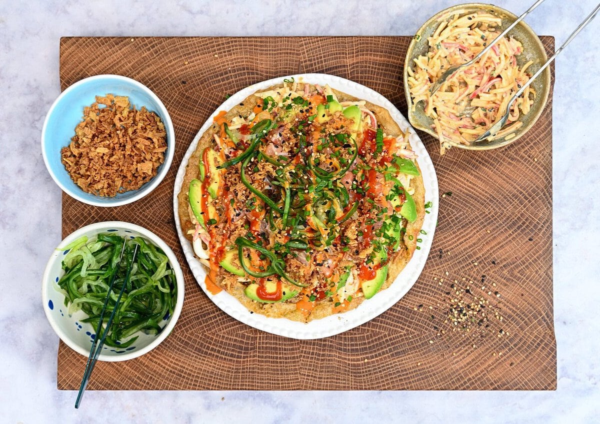 A plate of colorful sushi pizza topped with avocado, vegetables, and sesame seeds is on a wooden board. Surrounding it are bowls with crispy onions, cucumber strips, and a creamy slaw with tongs.