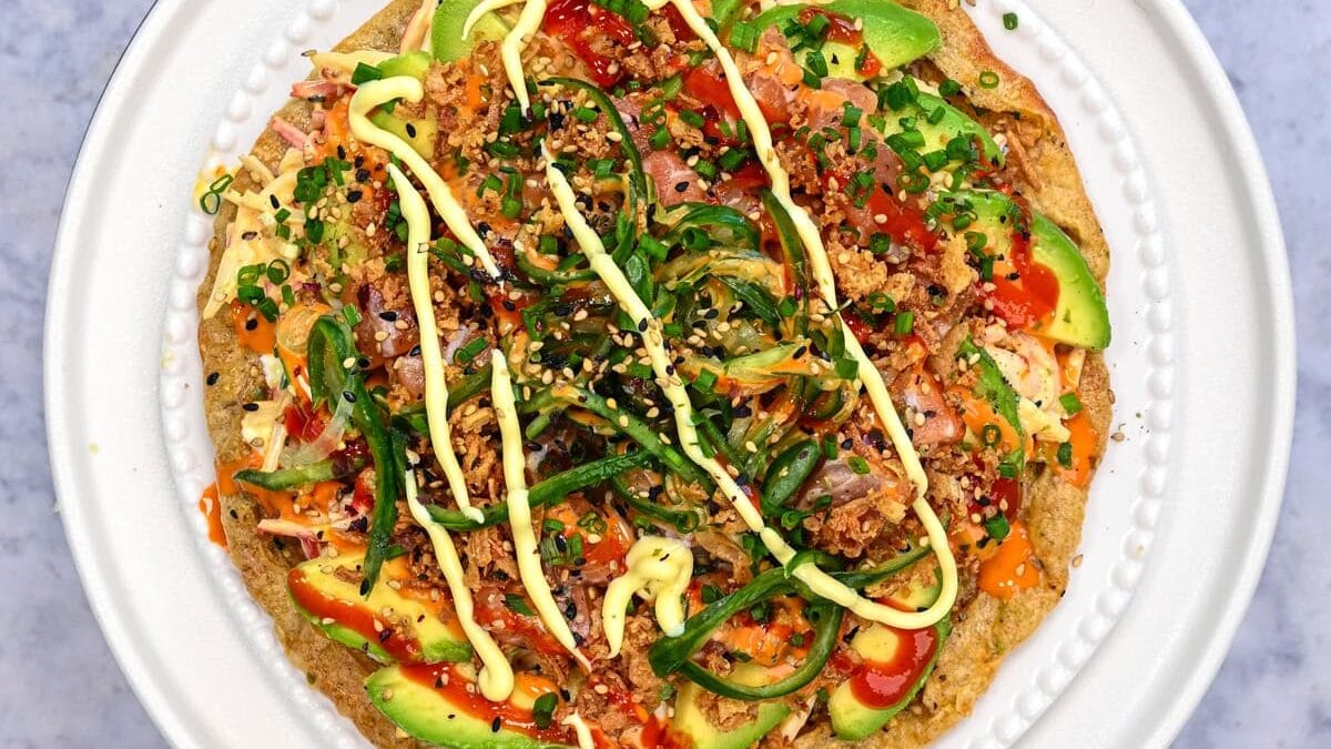 A colorful dish on a white plate features sliced avocado, greens, and a drizzle of yellow sauce, garnished with herbs and possibly quinoa or crab sticks, on a marble surface.