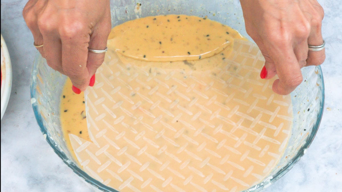 Two hands are dipping a circular sheet of rice paper into a bowl of light-colored, seasoned liquid, preparing it for use in cooking.
