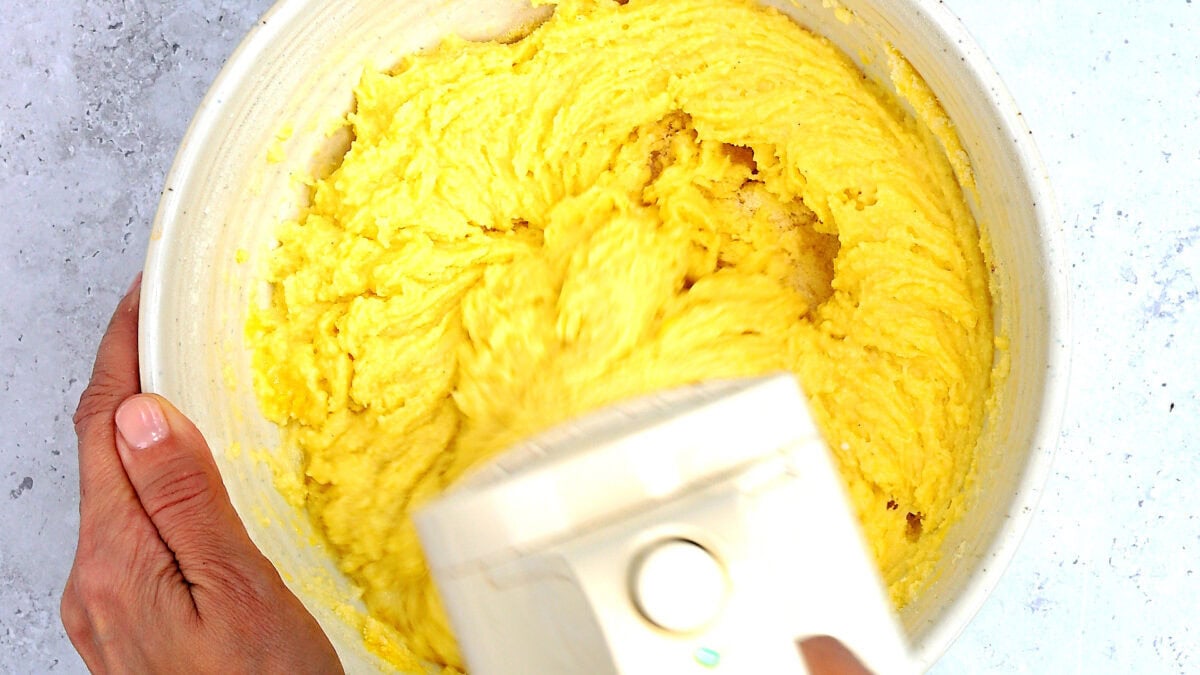 A person holds a bowl of yellow Whole Lemon Cake batter while mixing it with an electric hand mixer on a light surface.
