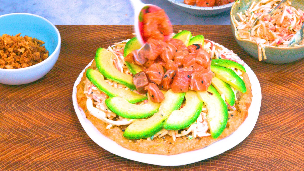 A tostada topped with shredded vegetables and sliced avocado is being garnished with cubed raw fish using a spoon. Bowls of toppings are visible in the background on a wooden surface.