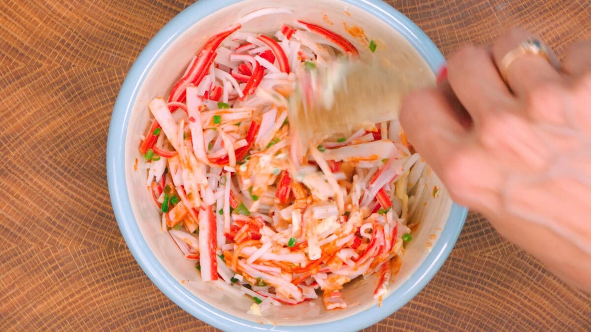 A hand is mixing a salad with imitation crab sticks, thinly sliced vegetables, and herbs in a white bowl on a wooden surface.