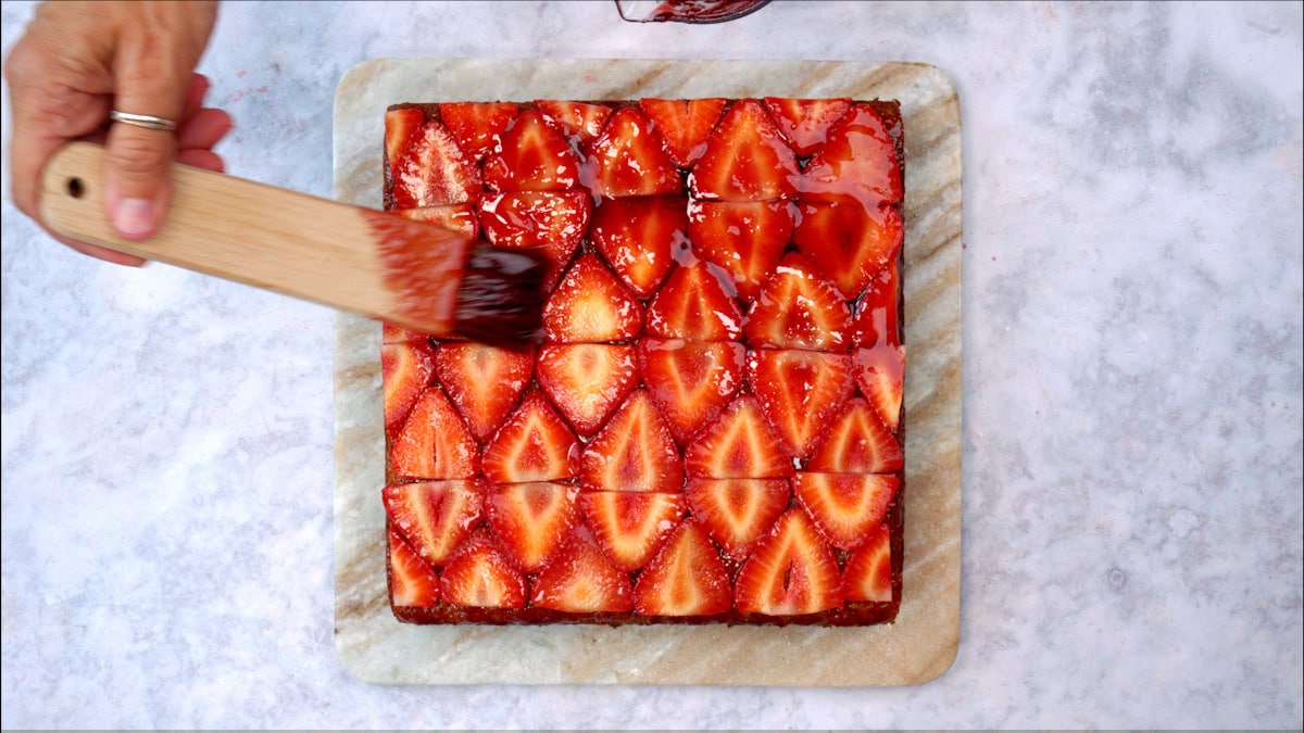 A hand uses a pastry brush to glaze a square strawberry tart cut into 16 pieces on a marble board.