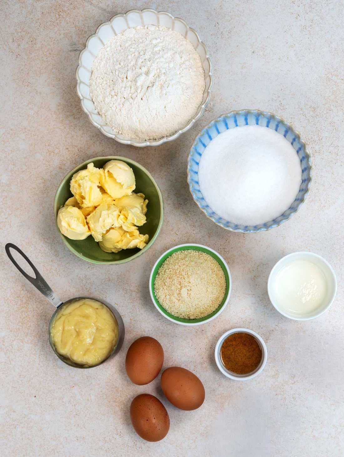Top view of baking ingredients: flour, sugar, butter, almond meal, milk, three eggs, blended boiled lemon paste, and a small bowl of ground spice, all arranged in separate bowls and measuring cups on a light surface.
