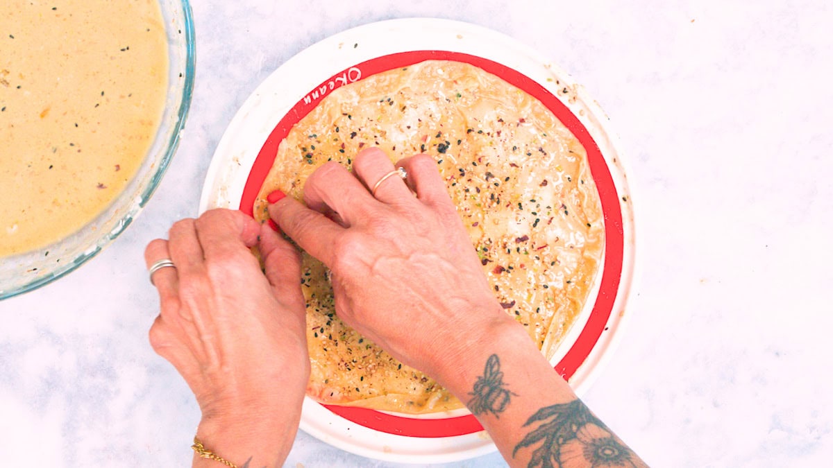 Hands folding a spiced flatbread on a red and white plate, with a bowl of batter nearby. One hand has rings and a bracelet, and a tattoo of flowers is visible on the wrist and forearm.