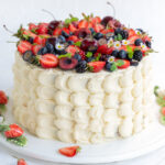 A tall, round Berry Chantilly Cake with white frosting decorated with piped scallops, topped with strawberries, blueberries, blackberries, cherries, small flowers, and fresh mint leaves. Some berries and leaves are scattered on the table.