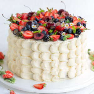 A tall, round Berry Chantilly Cake with white frosting decorated with piped scallops, topped with strawberries, blueberries, blackberries, cherries, small flowers, and fresh mint leaves. Some berries and leaves are scattered on the table.