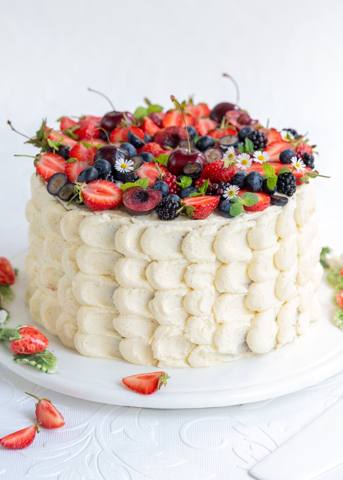 A tall, round Berry Chantilly Cake with white frosting decorated with piped scallops, topped with strawberries, blueberries, blackberries, cherries, small flowers, and fresh mint leaves. Some berries and leaves are scattered on the table.