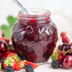 A glass jar filled with low sugar berry jam sits on a table surrounded by fresh strawberries, cherries, and blackberries, with a spoon inside the jar.