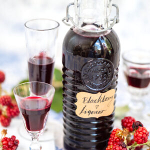 A bottle labeled Blackberry Liqueur sits among fresh blackberries, with two partially filled glasses nearby. The scene is bright and inviting, emphasizing the deep purple color of the drink—an irresistible reminder that you can make your own Blackberry Liqueur (Crème de Mûre) at home with just 4 simple ingredients. This rich, ruby-red liqueur is perfect for Bramble cocktails.