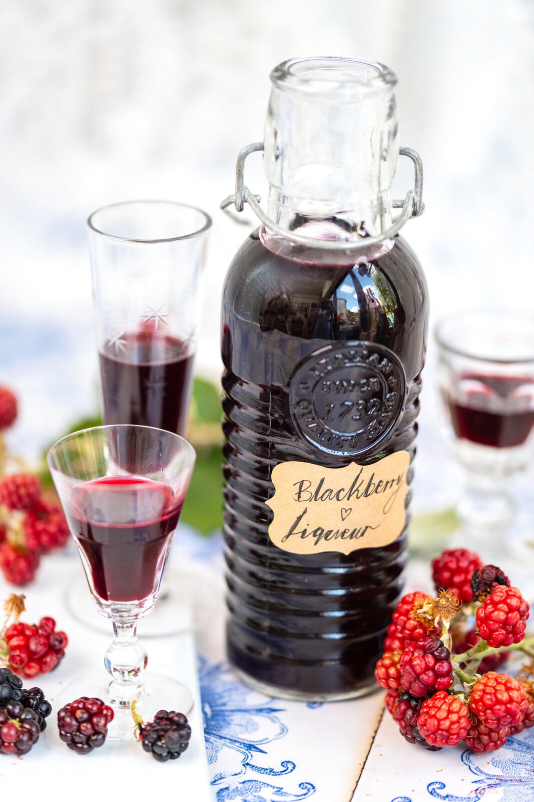 A bottle labeled Blackberry Liqueur surrounded by fresh blackberries and two glasses of liqueur, all set on a white and blue table.
