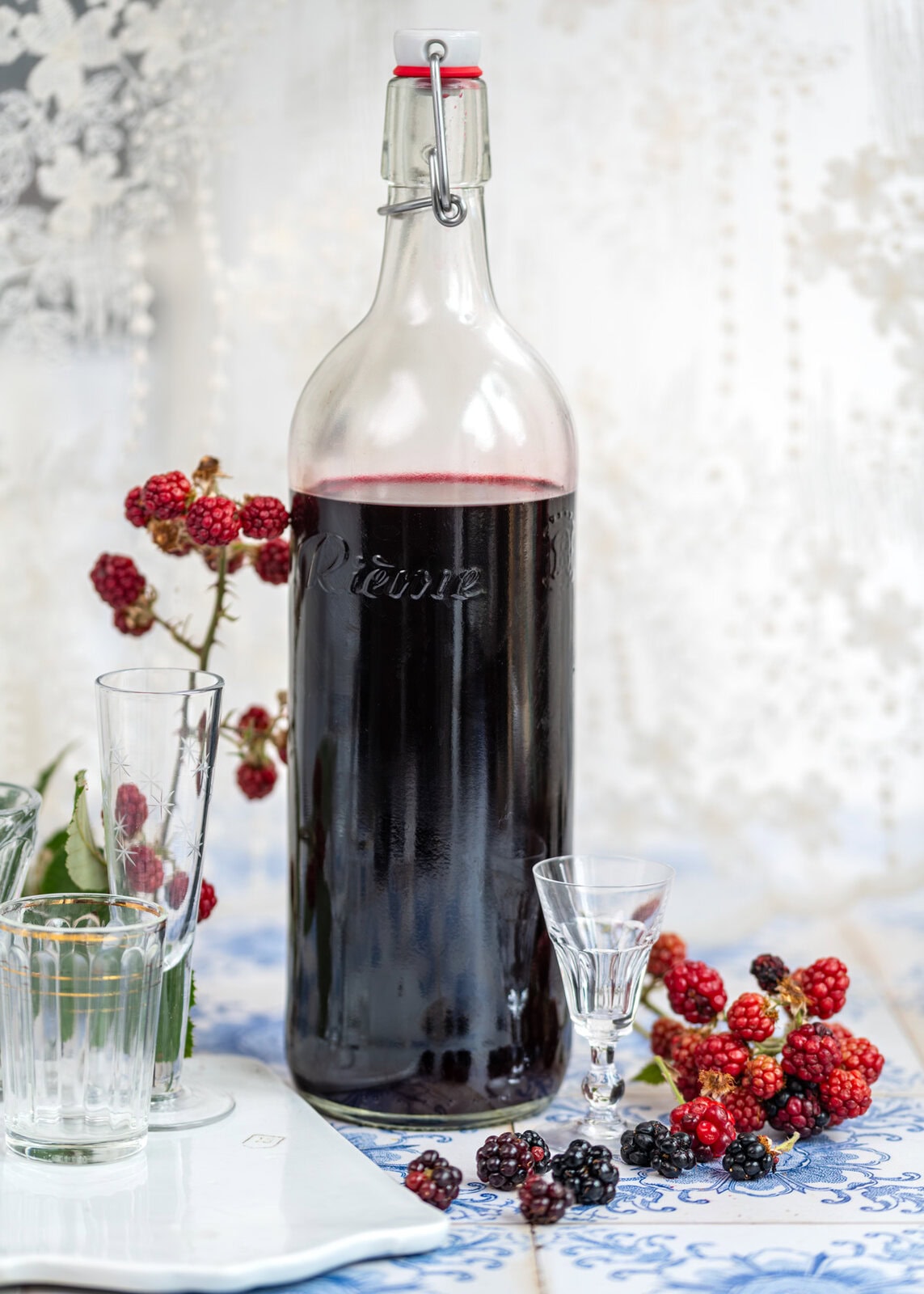 A tall glass bottle filled with dark liquid sits on a table with shot glasses, surrounded by ripe blackberries and blackberry branches.